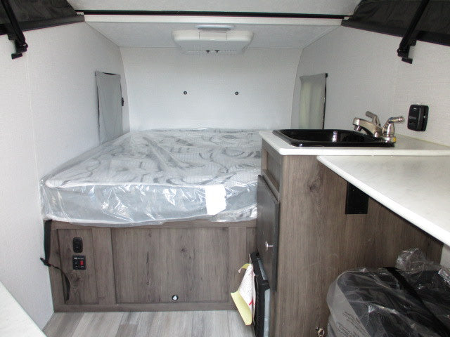 RV rental interior showcasing a bed with protective wrap, a compact kitchen with a sink, and modern wood-look finishes.