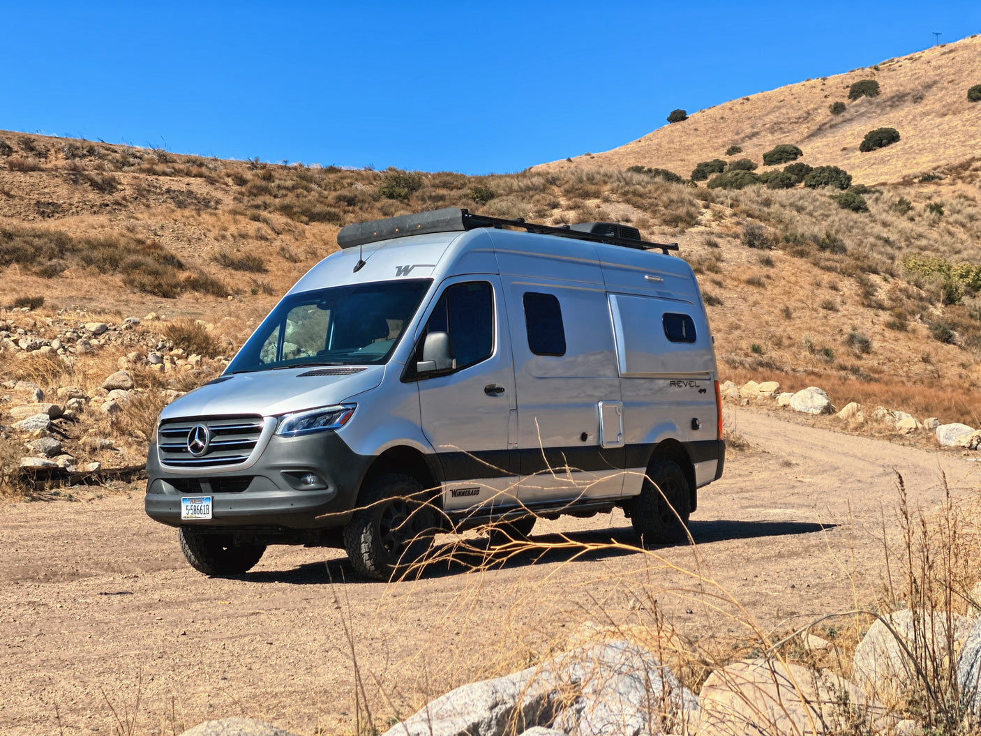 A silver Winnebago Revel rv rental, a camper van, is parked on a dirt road amidst dry, rolling hills.