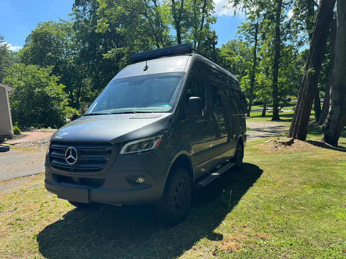 Dark green Mercedes-Benz Sprinter RV rental van parked on grass near a driveway & trees on a sunny day.
