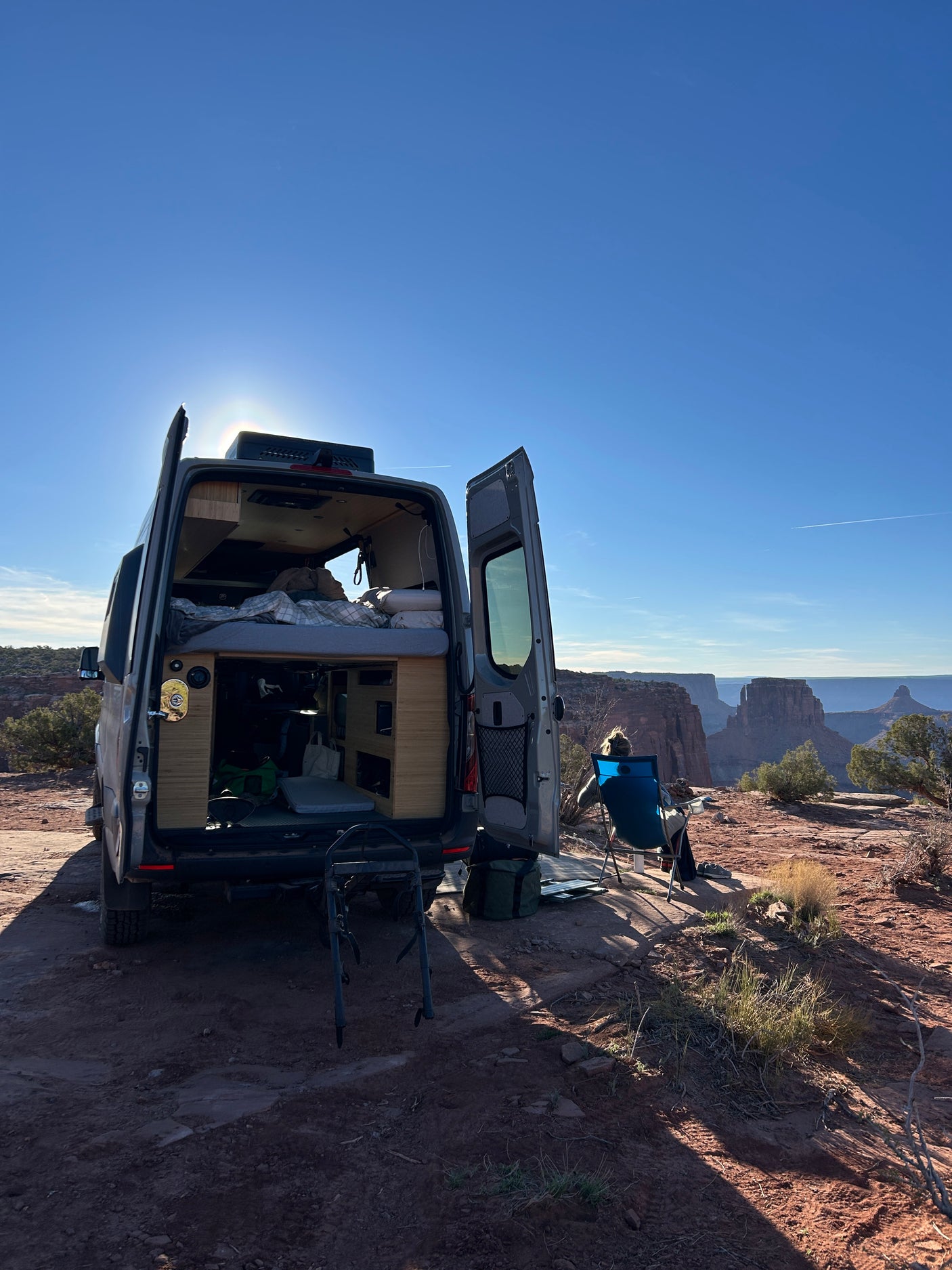 This RV rental provides a perfect view. A camper van is parked with open doors, revealing a cozy interior as a person enjoys the vast canyon vista.