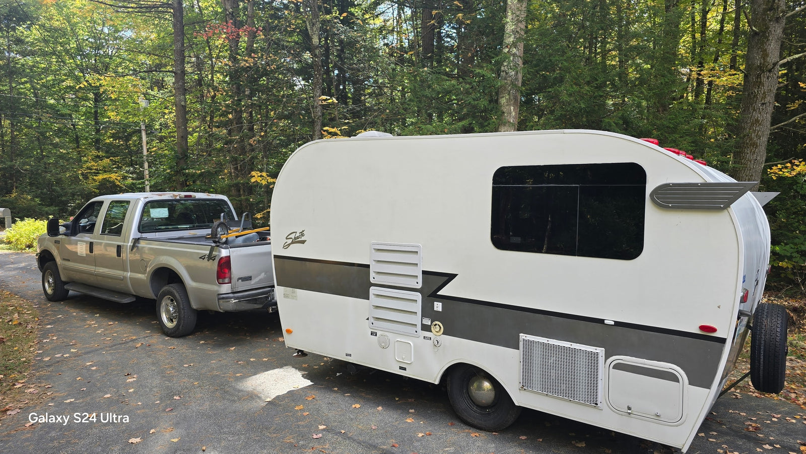 A silver pickup truck tows an RV rental on a leaf-covered road, set against a backdrop of colorful autumn trees. Perfect for outdoor travel!