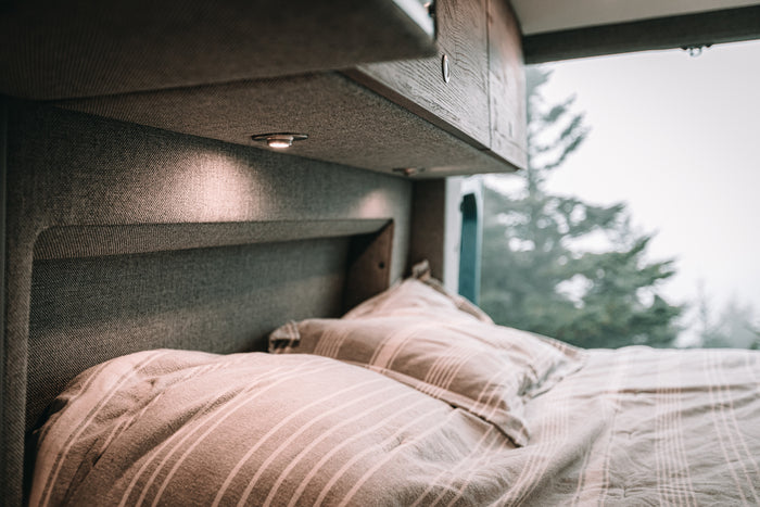 Cozy RV rental interior showing a comfortable bed with striped bedding, warm lighting, and a blurry view of misty evergreen trees outside.