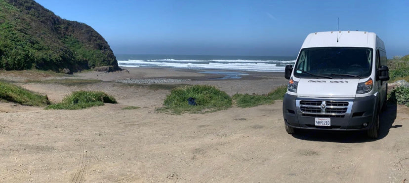 White RV rental van parked on a dirt road overlooking a scenic California beach with ocean waves. Perfect for coastal adventures.