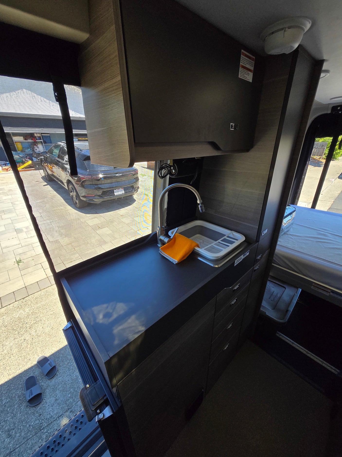 Compact RV rental interior featuring a kitchen sink, bed area, and an open side door view of an electric car outside.