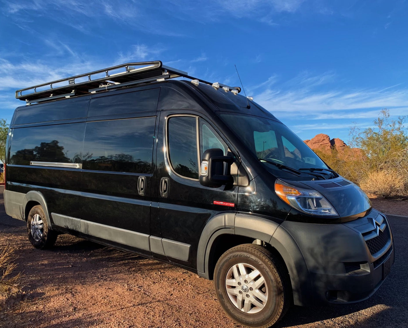 Black camper van RV rental with roof rack, on rugged terrain under blue sky, perfect for outdoor exploration.