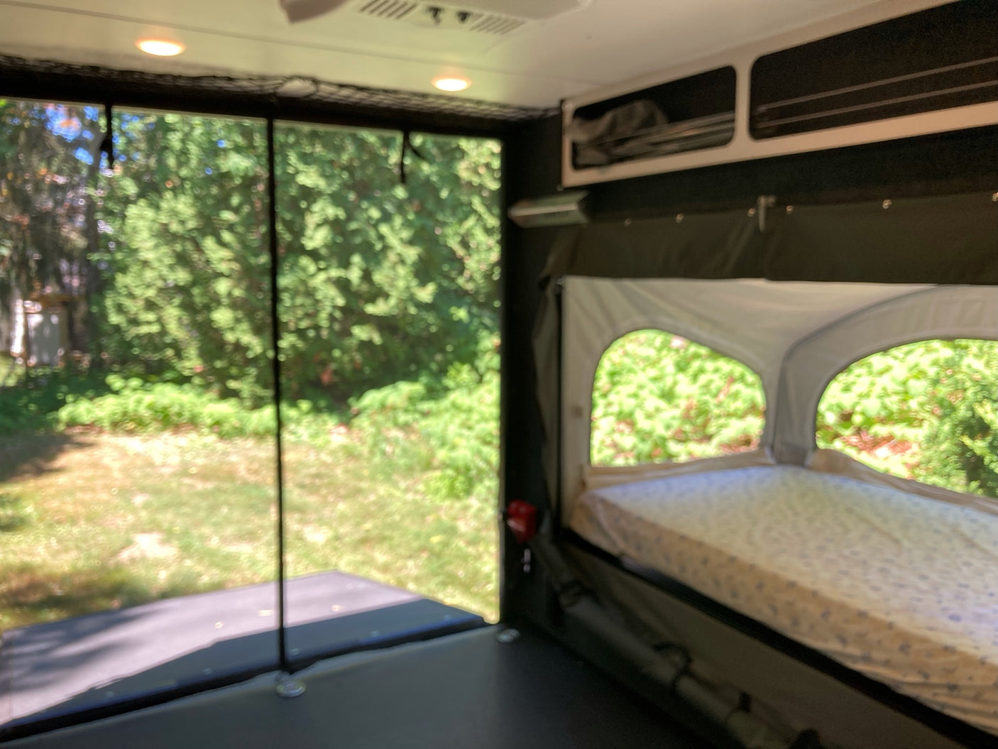 Interior of an RV rental: a pop-out bed on the right, large mesh screen on the left looking out to green trees and grass.