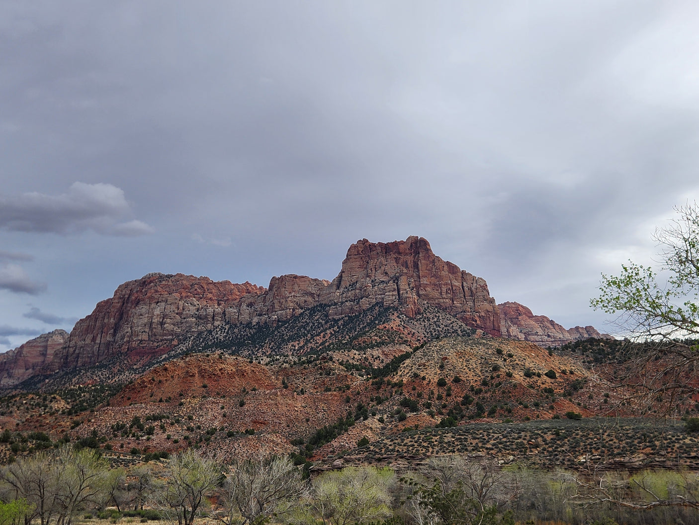 Explore this majestic red rock landscape with an RV rental, seeing impressive mountains and sparse desert flora under cloudy skies.
