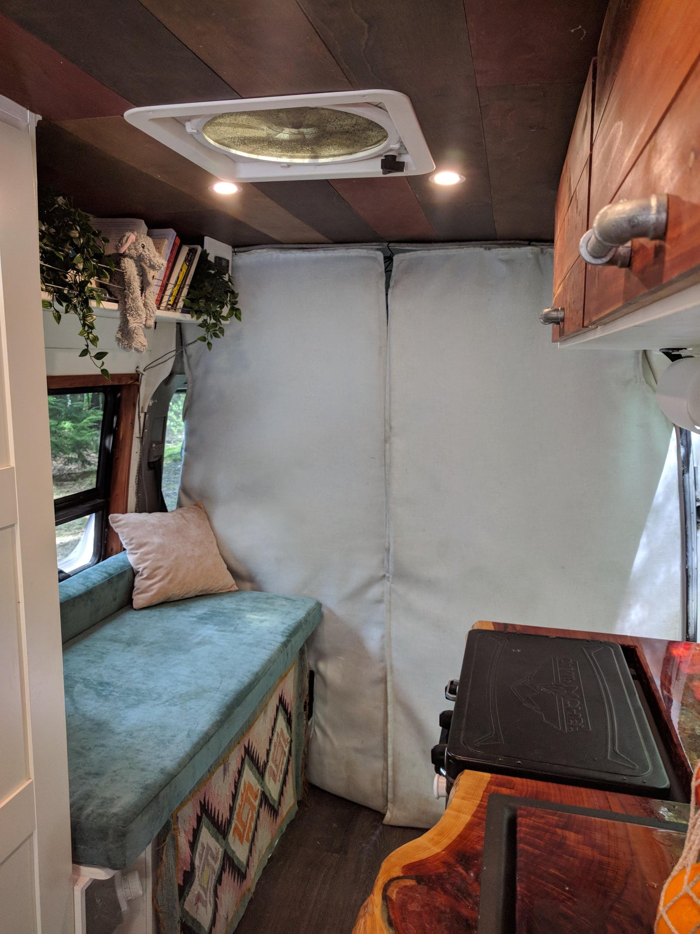 A cozy rv rental interior features a teal cushioned bench, wood-paneled ceiling with a fan, and a compact kitchen setup.