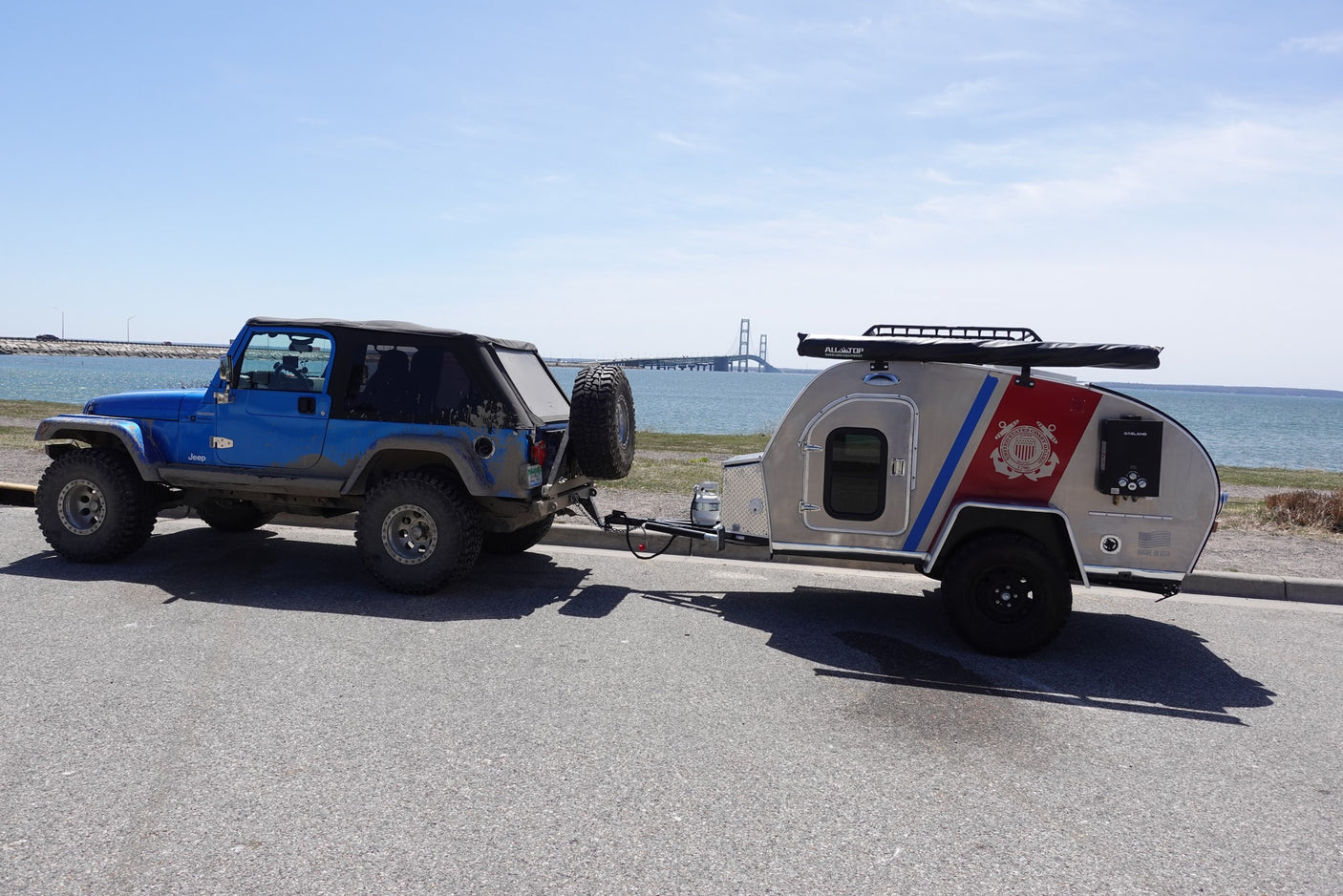 Overland RV rental: Blue Jeep towing a unique Coast Guard teardrop trailer by a scenic bridge.