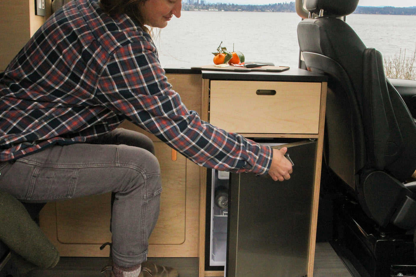 Exploring van life: a person in a plaid shirt opens the refrigerator in their RV rental camper van kitchenette by a lake.