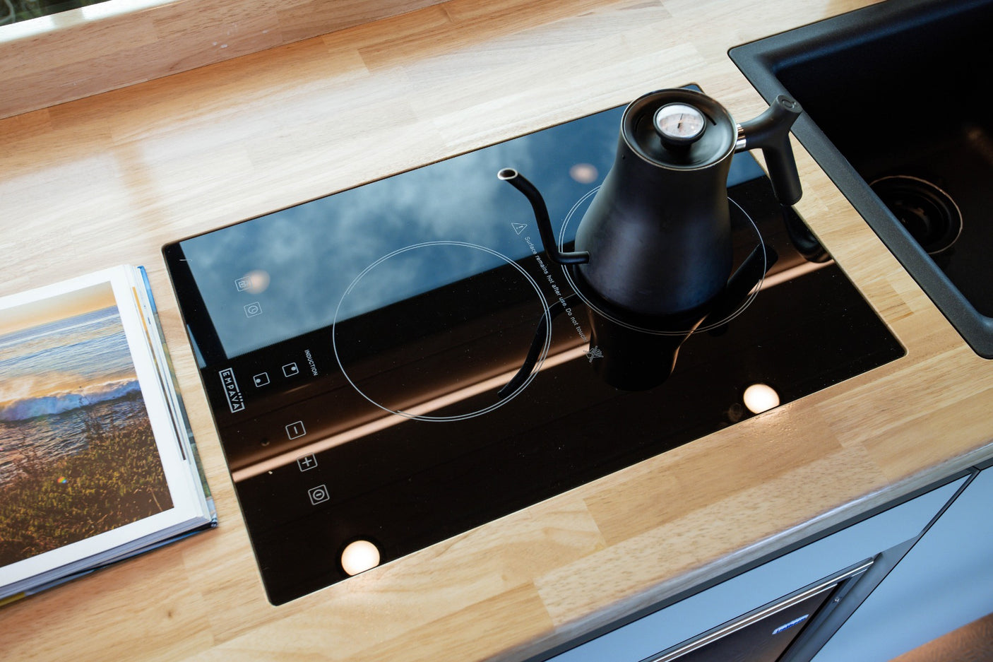 Compact RV rental kitchen features a sleek induction cooktop with a kettle on a light wood counter.