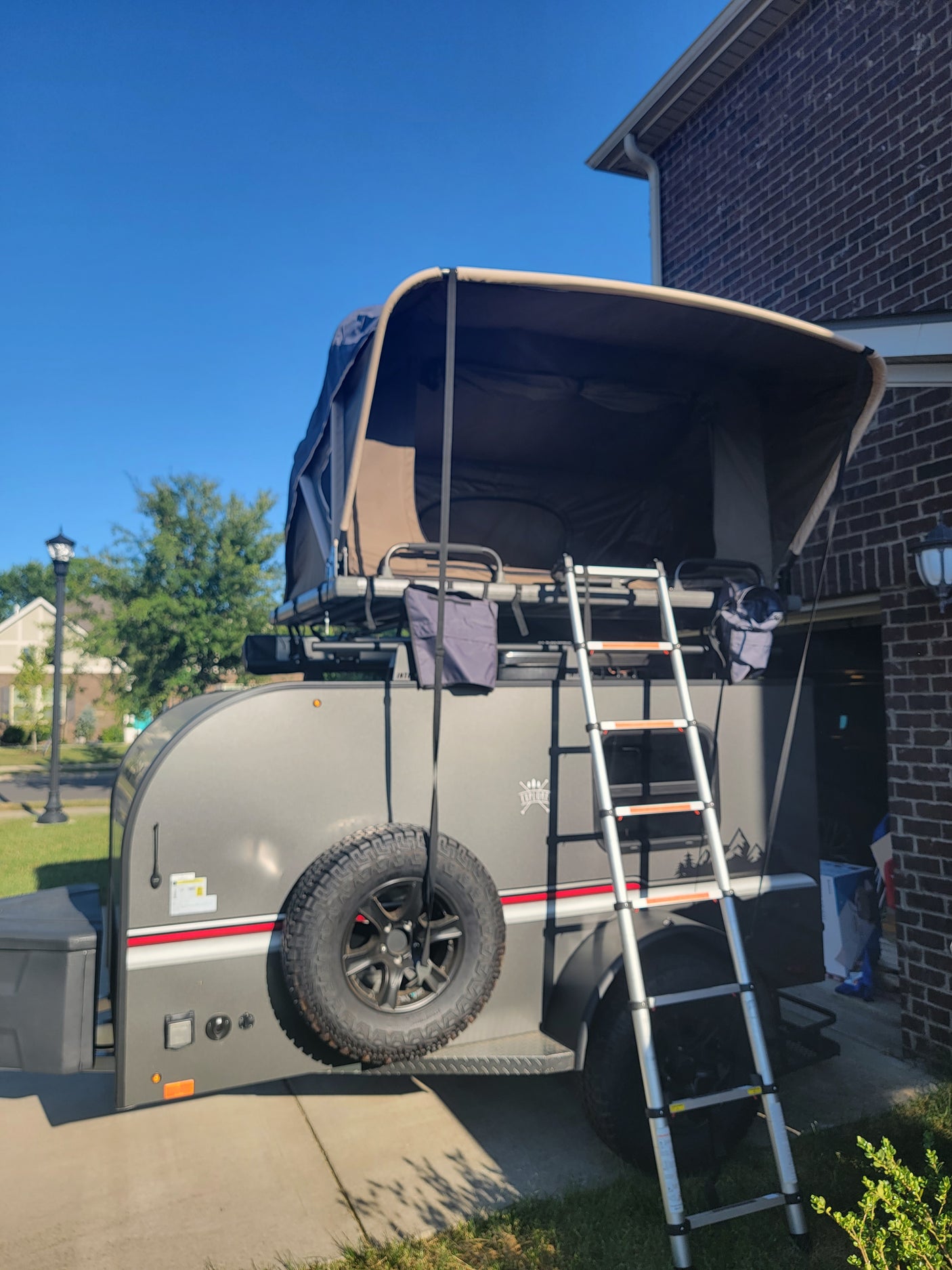 Grey pop-up camper for RV rental, featuring ladder & spare tire, parked on a sunny driveway.