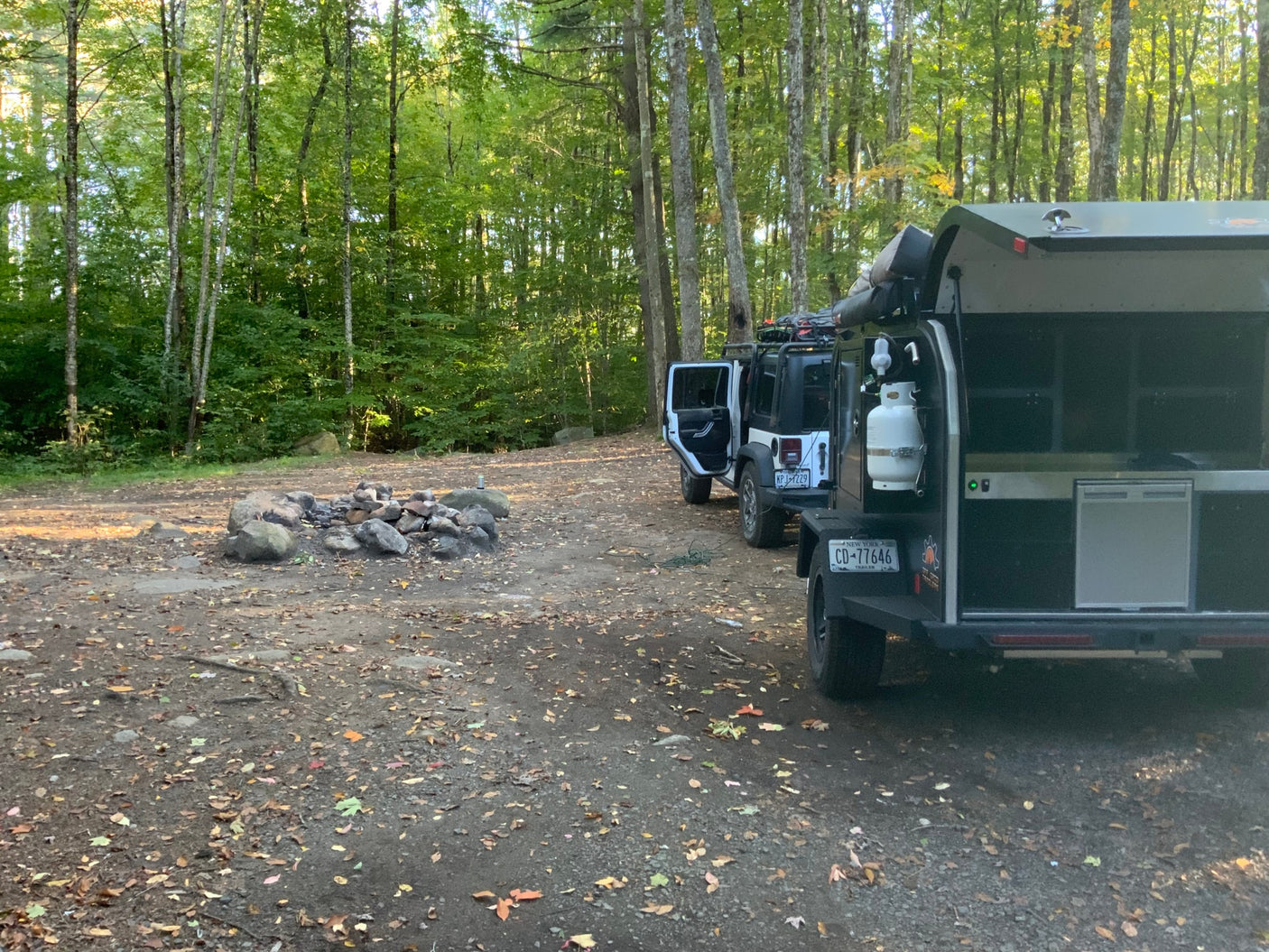 ready for outdoor cooking beside a Jeep.|White Jeep & overland RV rental trailer setup with portable solar panels at an autumn campsite amidst golden leaves.|A white Jeep and black overland trailer parked at a sunny campsite with a rock fire pit. Perfect for an RV rental outdoor adventure.|Off-grid rv rental camping setup with Jeep & overland trailer in a serene forest campsite