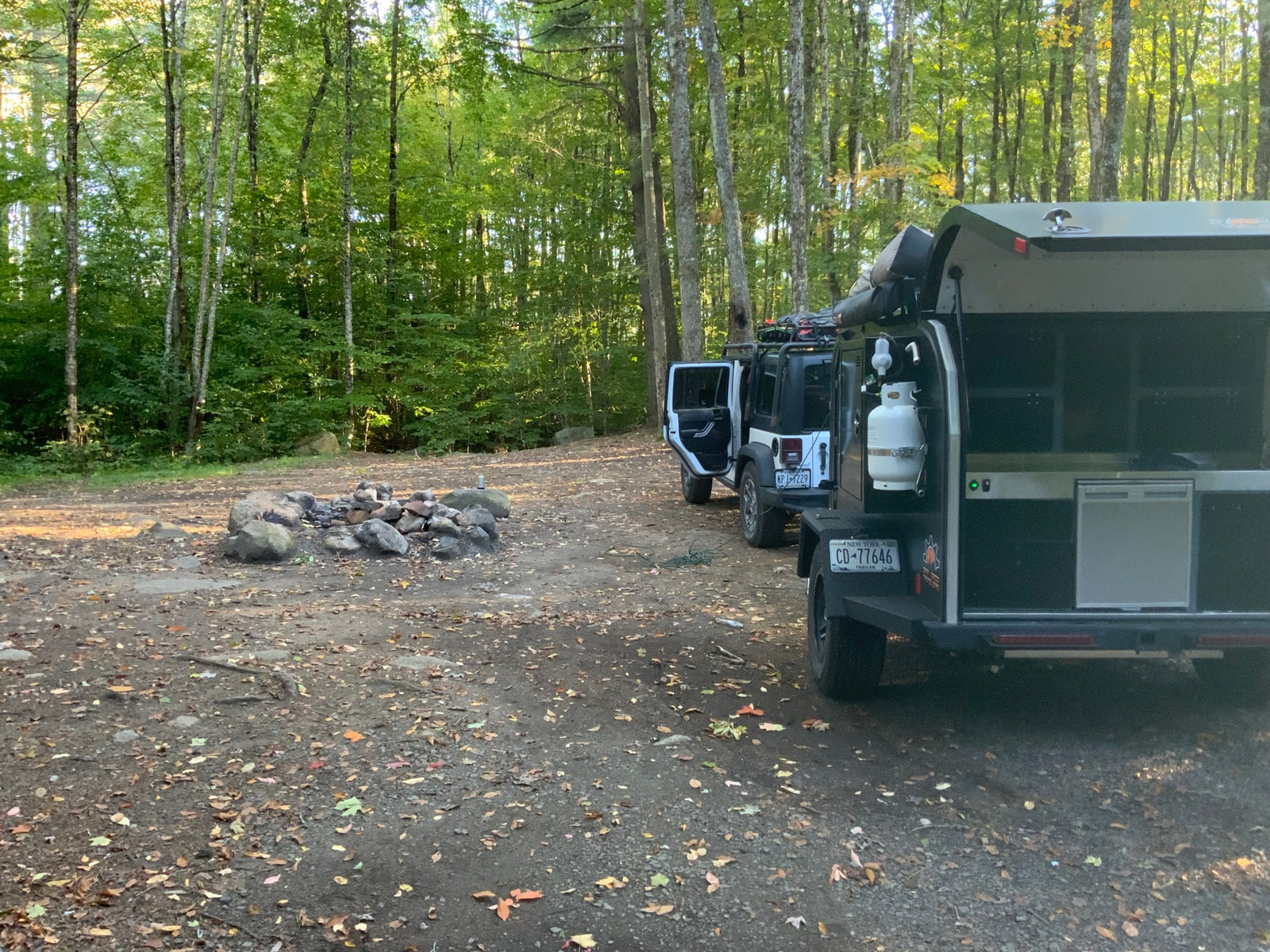 ready for outdoor cooking beside a Jeep.|White Jeep & overland RV rental trailer setup with portable solar panels at an autumn campsite amidst golden leaves.|A white Jeep and black overland trailer parked at a sunny campsite with a rock fire pit. Perfect for an RV rental outdoor adventure.|Off-grid rv rental camping setup with Jeep & overland trailer in a serene forest campsite