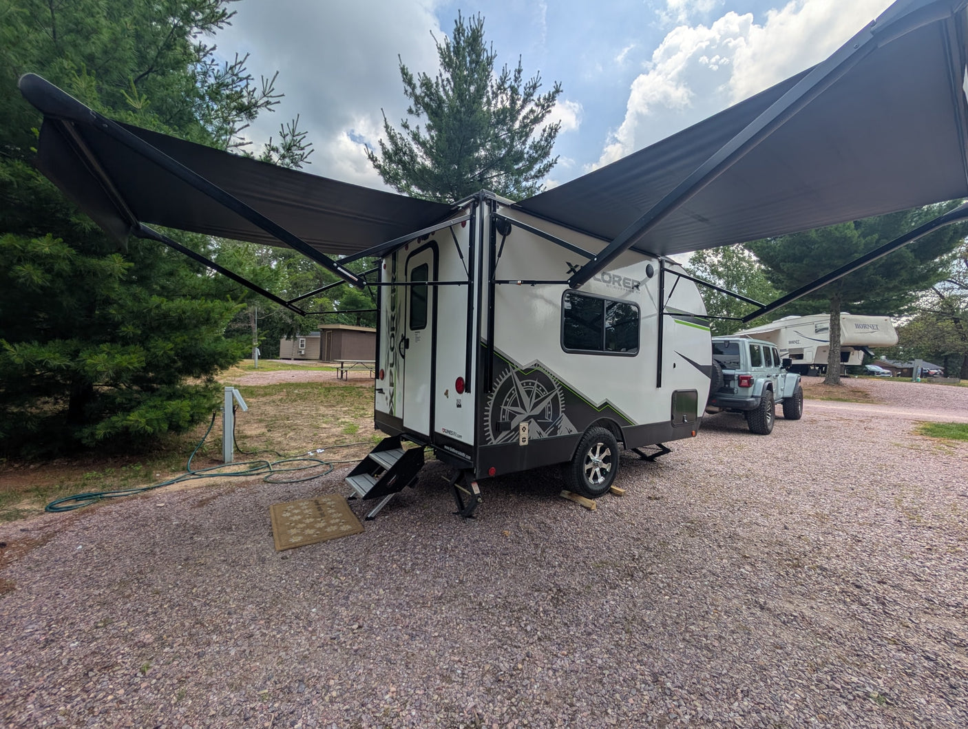 This RV rental, a white travel trailer with an extended awning, sits at a gravel campsite. A Jeep is parked nearby.