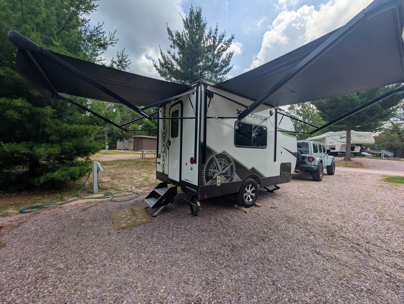 This RV rental, a white travel trailer with an extended awning, sits at a gravel campsite. A Jeep is parked nearby.