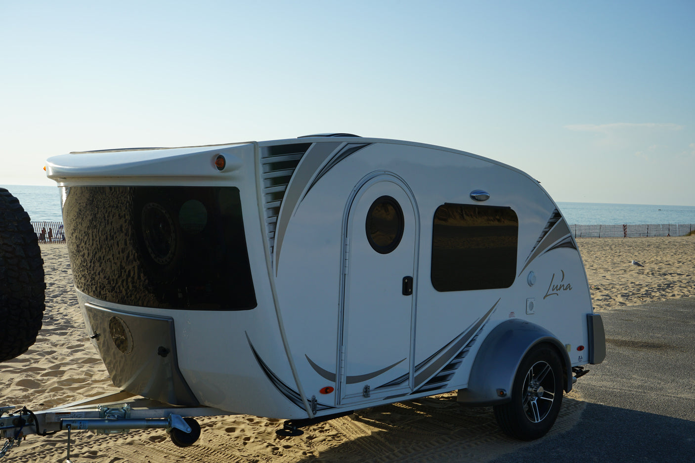 Teardrop RV rental parked on an asphalt path next to a sandy beach and ocean under a clear sky.