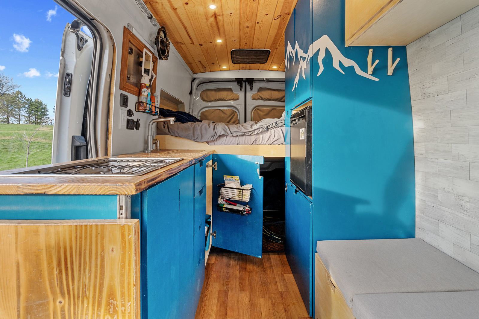 Inside this vibrant camper van RV rental: kitchen, bed, custom blue cabinetry with mountain art, and nature view.
