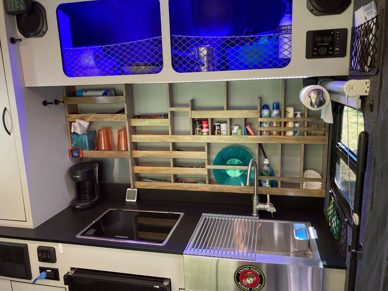 This modern RV rental galley features an induction cooktop, sink, organized shelving, and blue-lit overhead cabinets for efficient space.