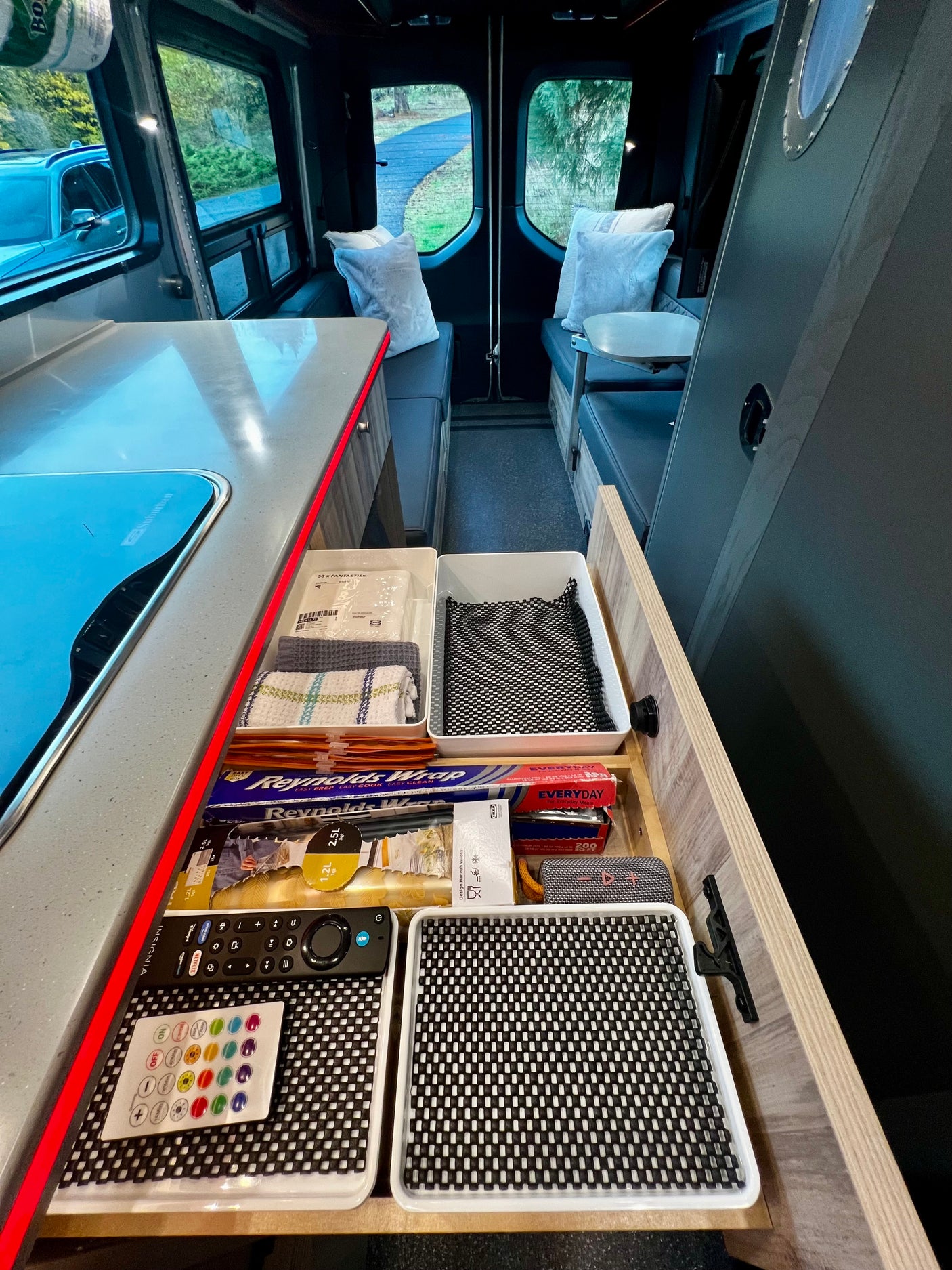 Organized kitchen drawer of an RV rental, featuring cooking supplies and remotes. A cozy living area is visible in the background.