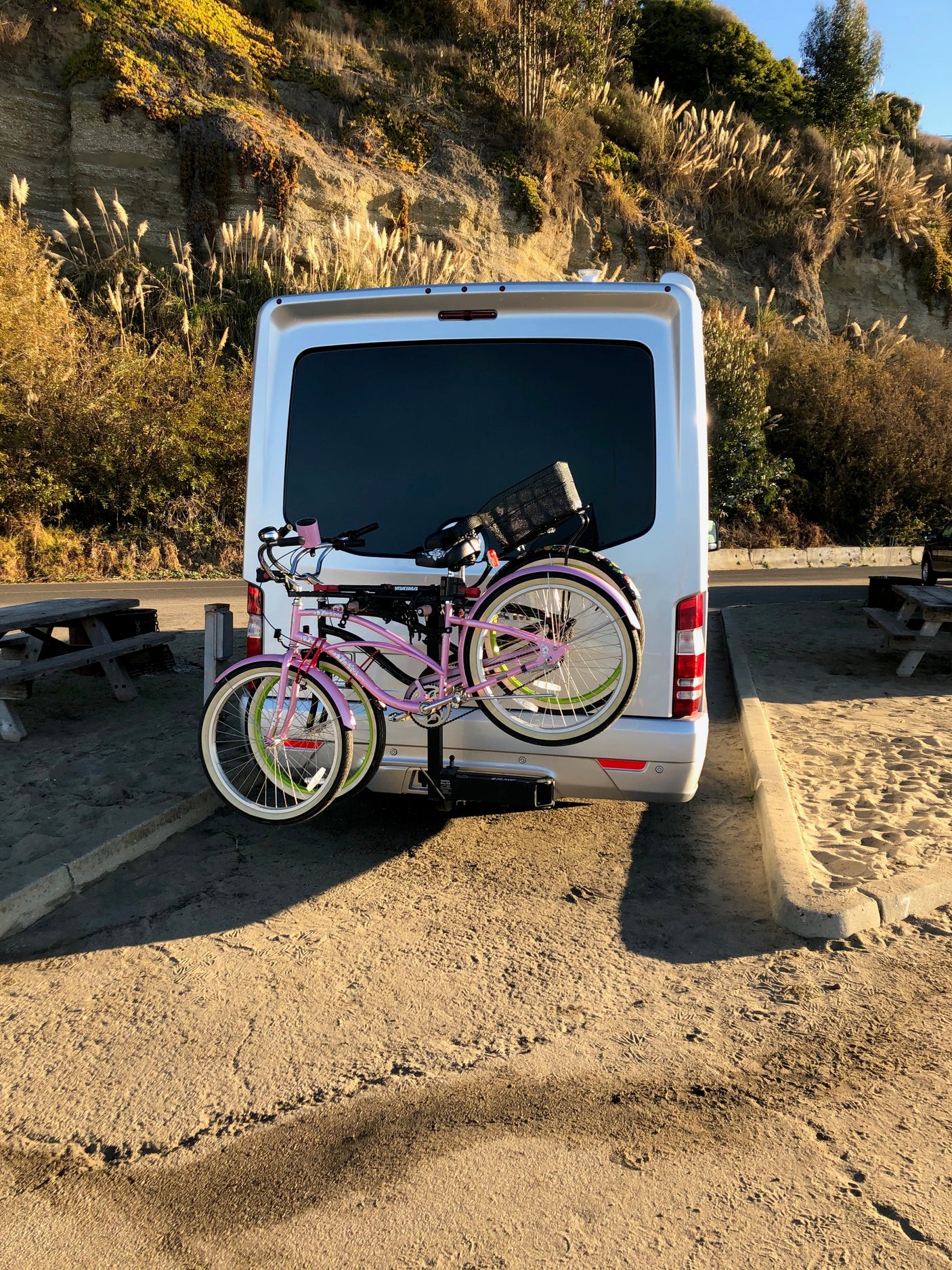An RV rental van carries bikes on its rear rack, parked by a sunlit sandy cliff. Ready for scenic outdoor adventures.