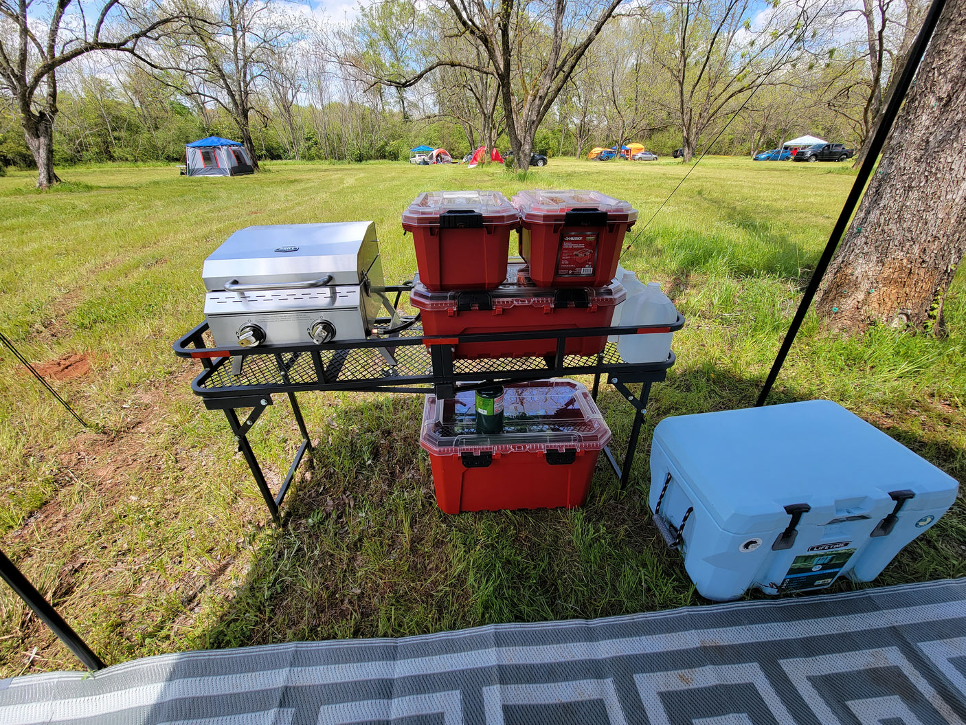 Portable camp kitchen with grill, red storage containers, and a blue cooler set up on a grassy field. Great for RV rental trips.