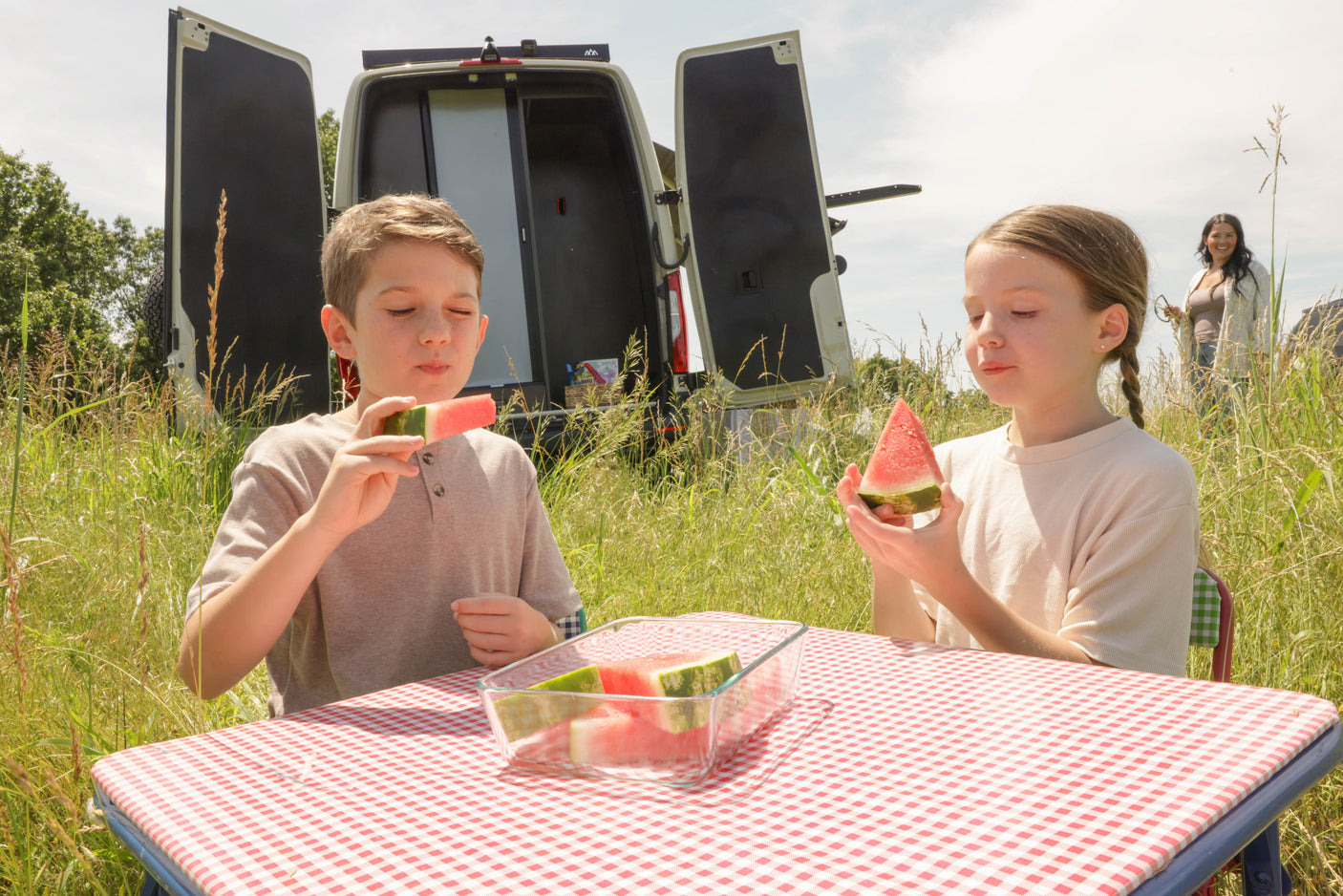 Two children enjoy watermelon at a picnic table, with an RV rental parked in a sunny, grassy field. An adult stands nearby.