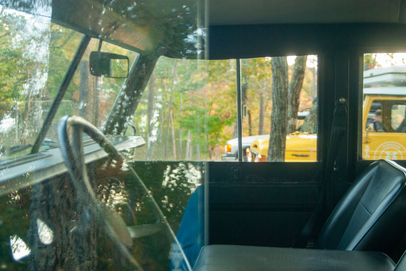 From inside a vintage Land Rover, viewing other yellow RV rentals and Land Rovers parked among fall trees in a forest.