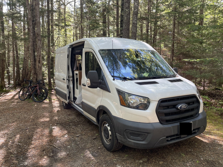 White Ford Transit camper van in a sunny forest with bikes. Your next RV rental adventure awaits!|RV rental: Cozy camper van interior with an elevated bed