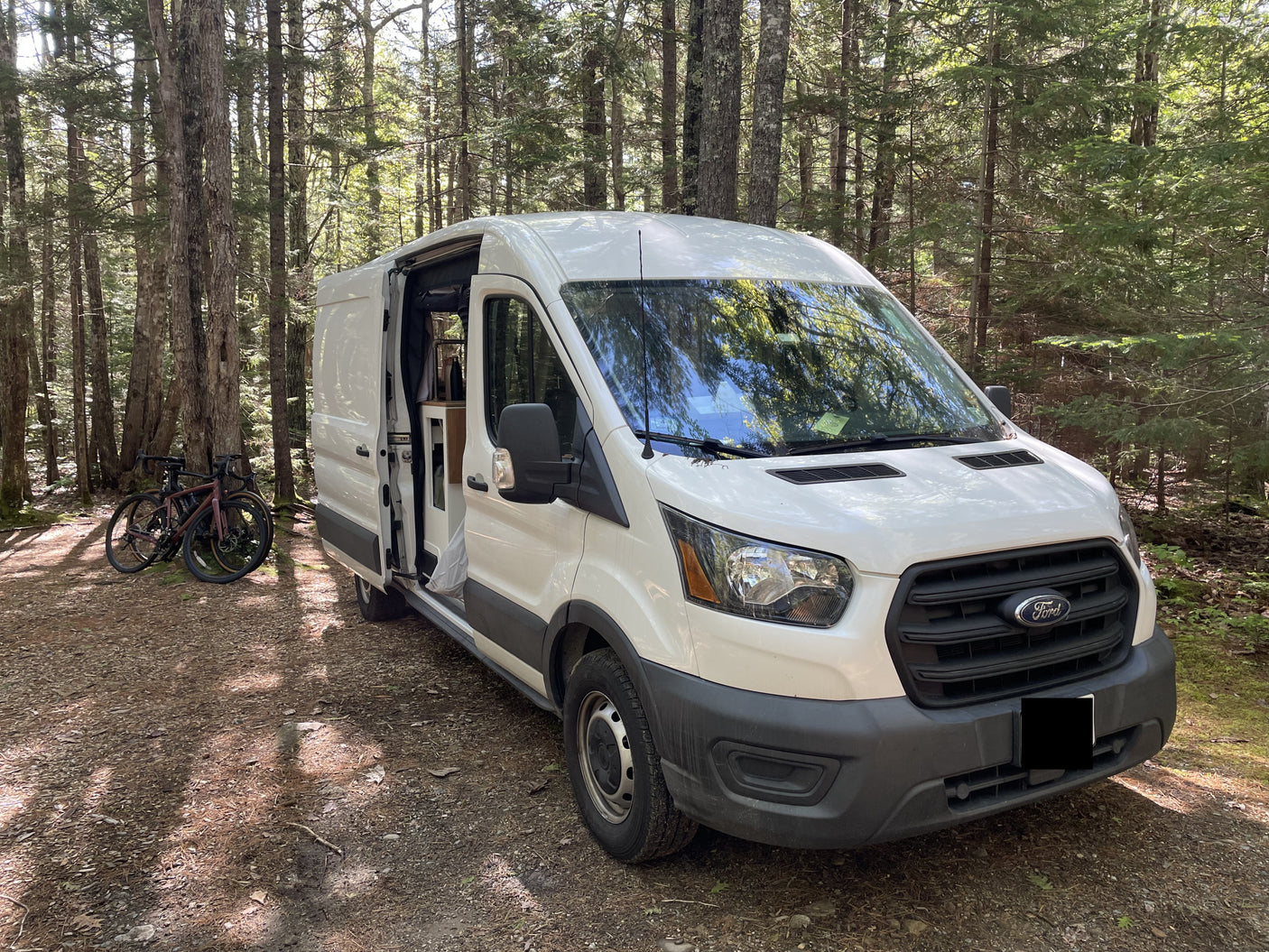 White Ford Transit camper van in a sunny forest with bikes. Your next RV rental adventure awaits!|RV rental: Cozy camper van interior with an elevated bed
