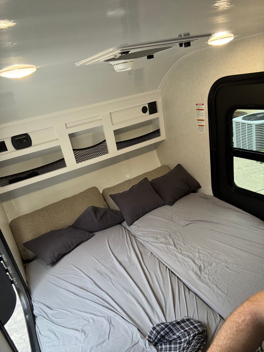 Cozy RV rental interior featuring a bedroom with gray bedding, overhead storage, charging center, ceiling lights, and window.