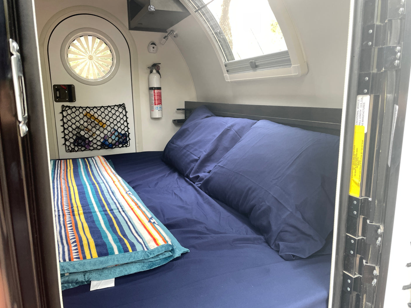 Cozy rv rental bedroom with a comfy blue bed, colorful blanket, windows, and a fire extinguisher for safety.