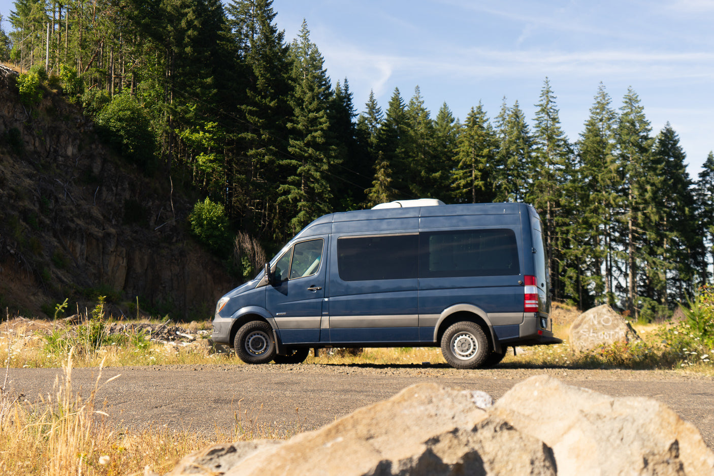 Blue RV rental van parked on a sunny road under a blue sky, with a dense forest and hillside behind it.