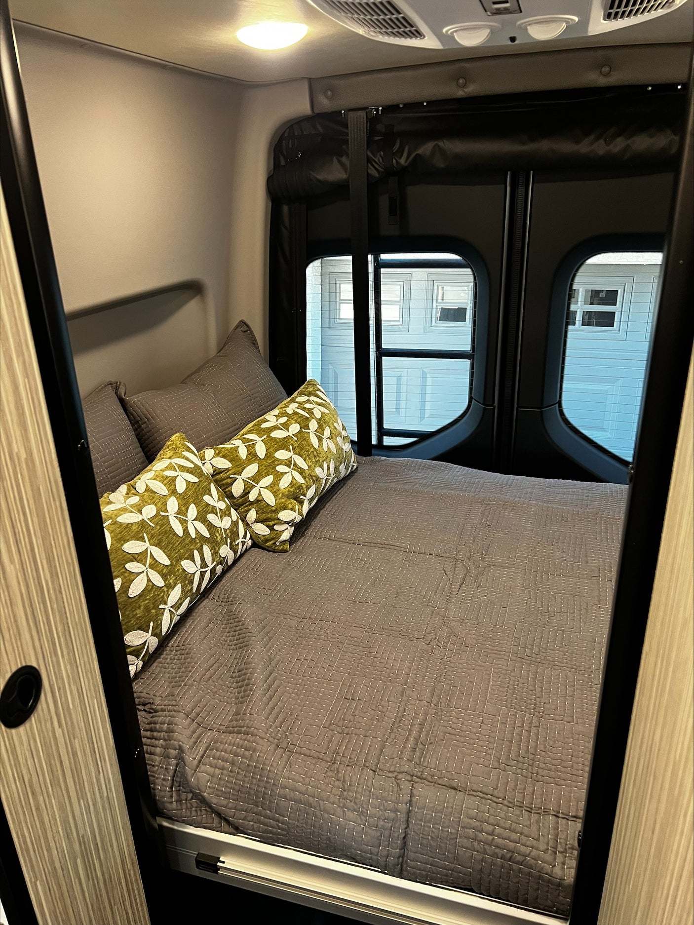 Cozy RV rental bedroom with a comfy bed, gray bedding, and green leaf-patterned pillows near small rear windows.
