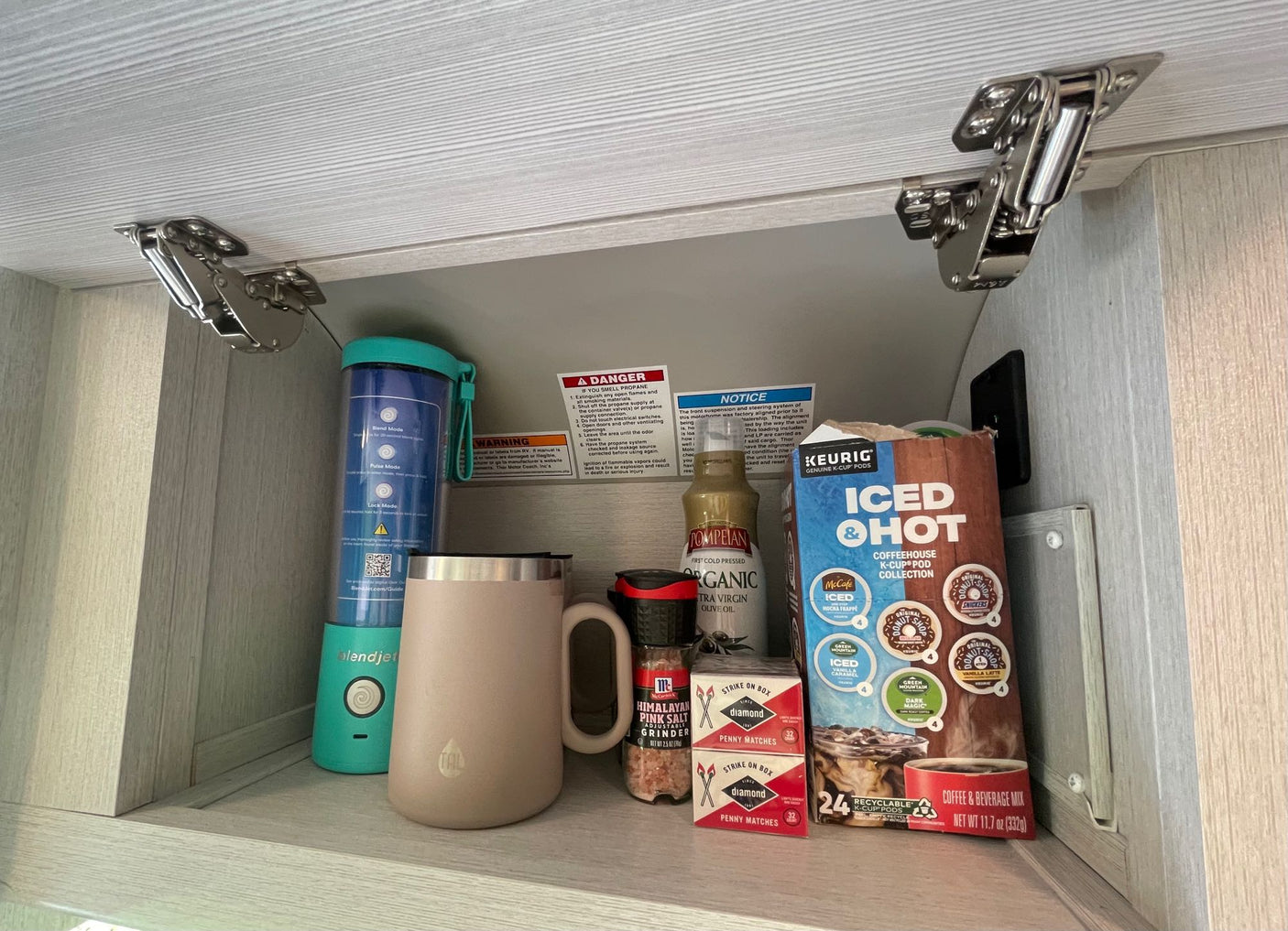 Inside an RV rental cabinet: a BlendJet, coffee pods, salt grinder, olive oil, and matches – ready for travel.