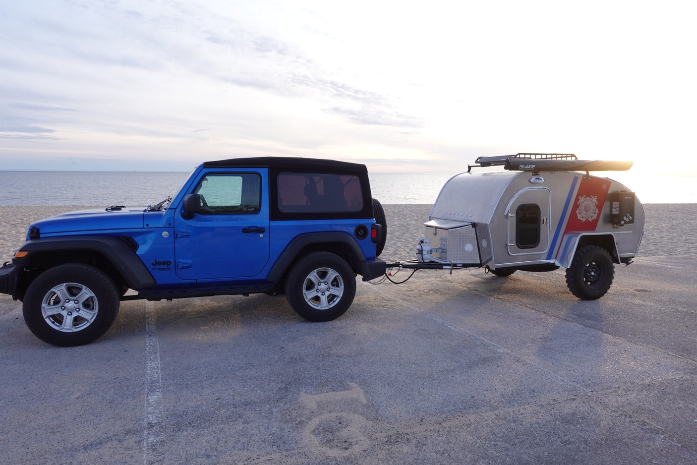 Blue Jeep Wrangler towing a silver travel trailer on concrete by a sandy beach and ocean. Great for an rv rental!