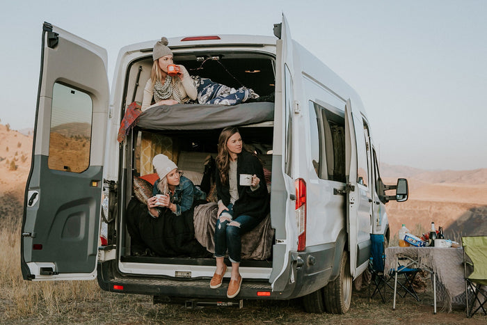 Three women relax in their RV rental campervan, enjoying drinks from the open back. A cozy van life scene amidst scenic hills.