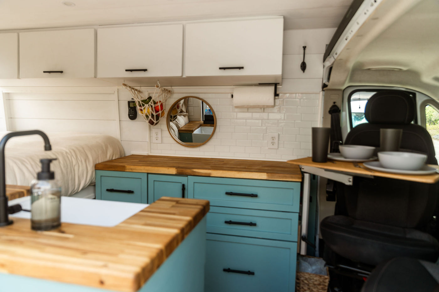 and a cozy bed.|White RV rental van parked by the ocean under tall trees. Its open side reveals a customized interior with light blue cabinets. Ideal for van life.|This cozy RV rental interior features a modern kitchen with blue cabinets — photo 3