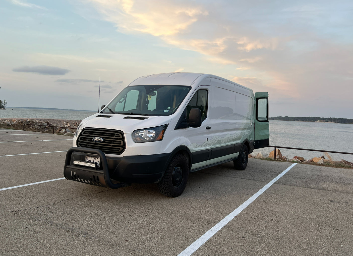 An RV rental ready for adventure: white Ford Transit camper van parked lakeside at dusk with the back door open, perfect for van life.