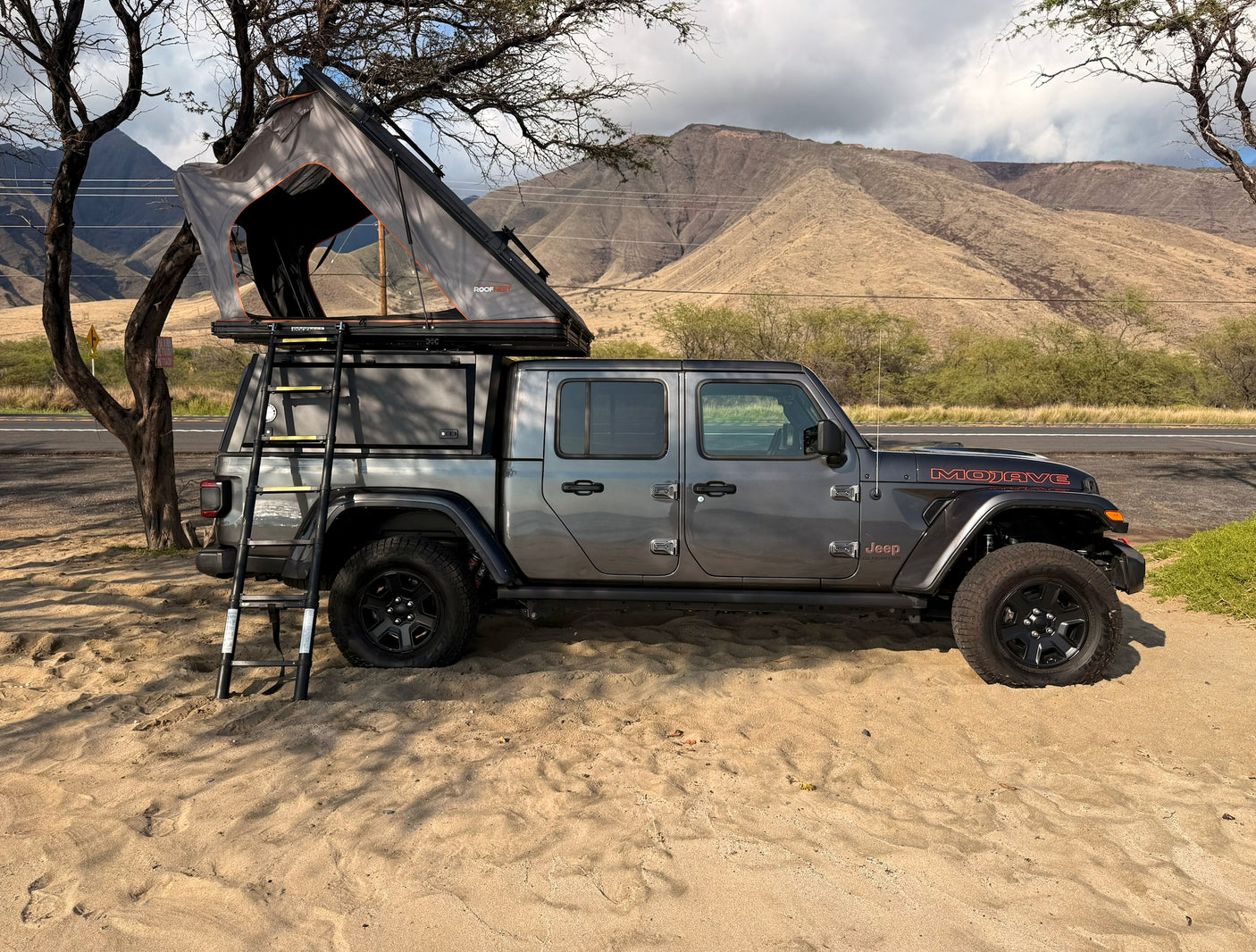 Experience outdoor adventure! This gray Jeep Gladiator, an ideal rv rental, has a roof-top tent & ladder, parked by mountains.