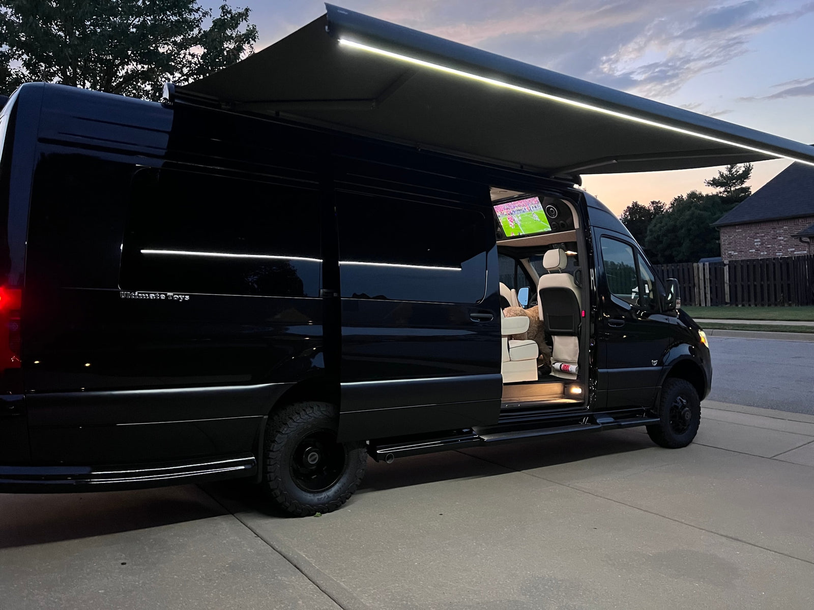 Luxury black Sprinter van with lit awning and interior TV showing sports; an excellent rv rental option.|A black Sprinter van