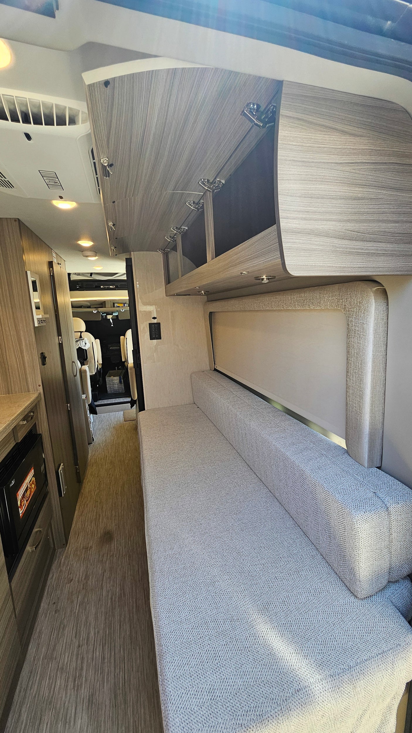 Modern RV rental interior with light wood cabinetry, a long grey fabric bench/bed, bright lighting, and vinyl flooring.