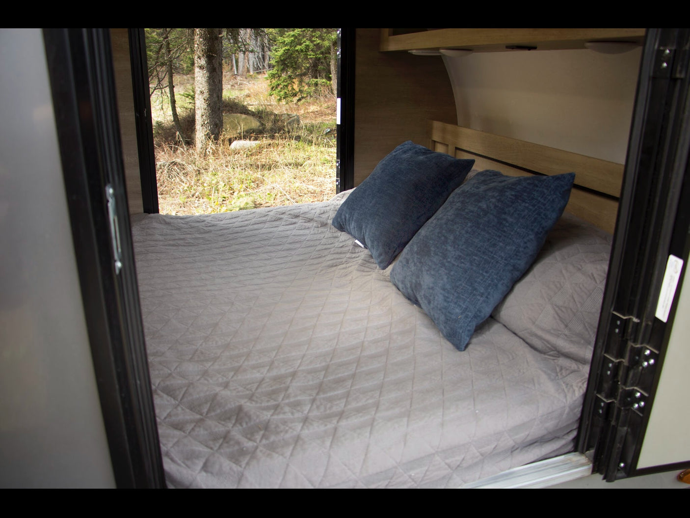 Inside this inviting RV rental: a comfortable bed with blue pillows and a large window overlooking a peaceful forest.