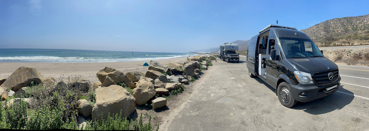 Coastal RV rental parked with an open door next to a sandy beach. Waves, other RVs, and hills complete the sunny scene.