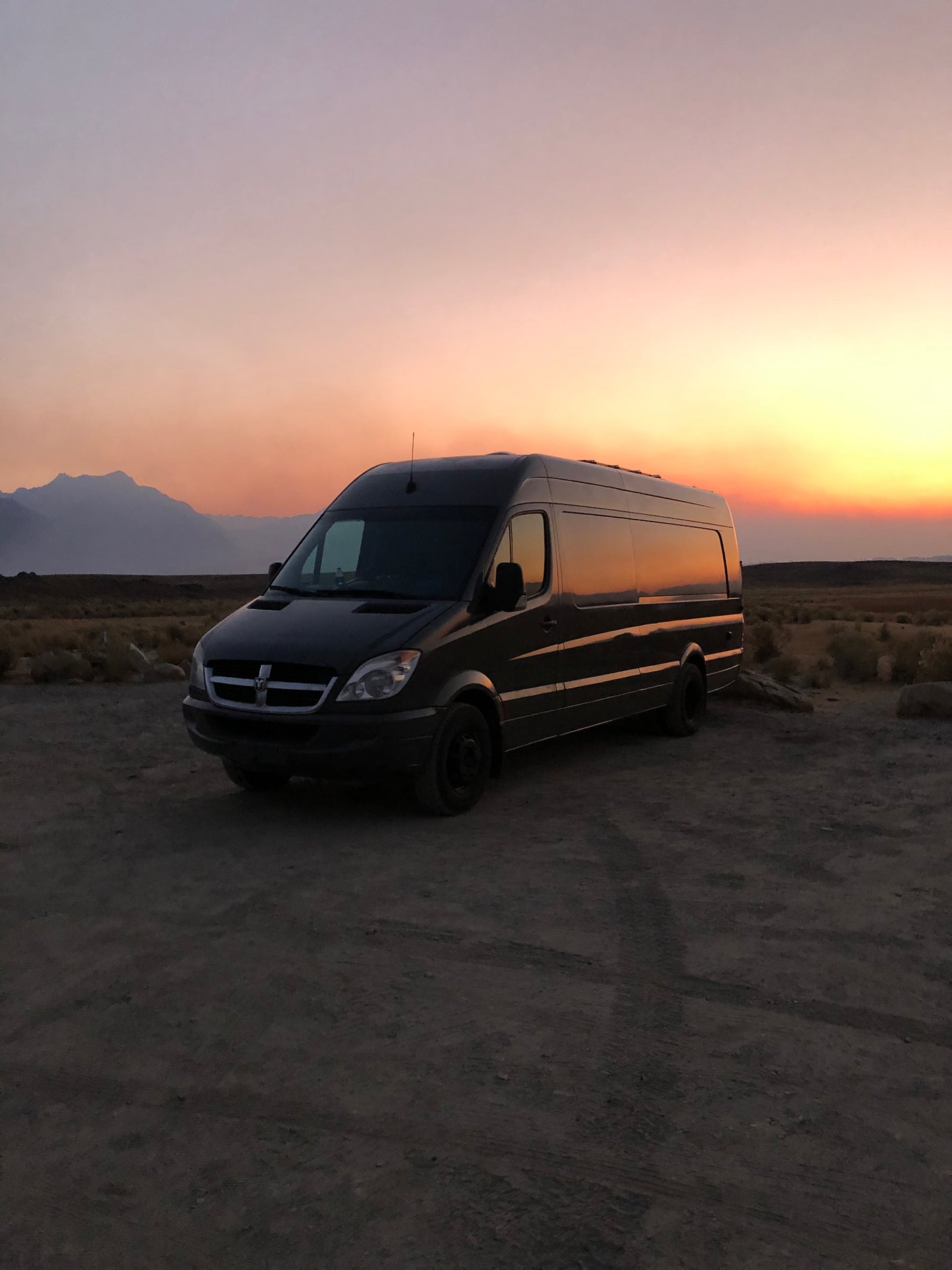 Dark Dodge Sprinter RV rental van silhouetted against a vibrant desert sunset with distant mountains.