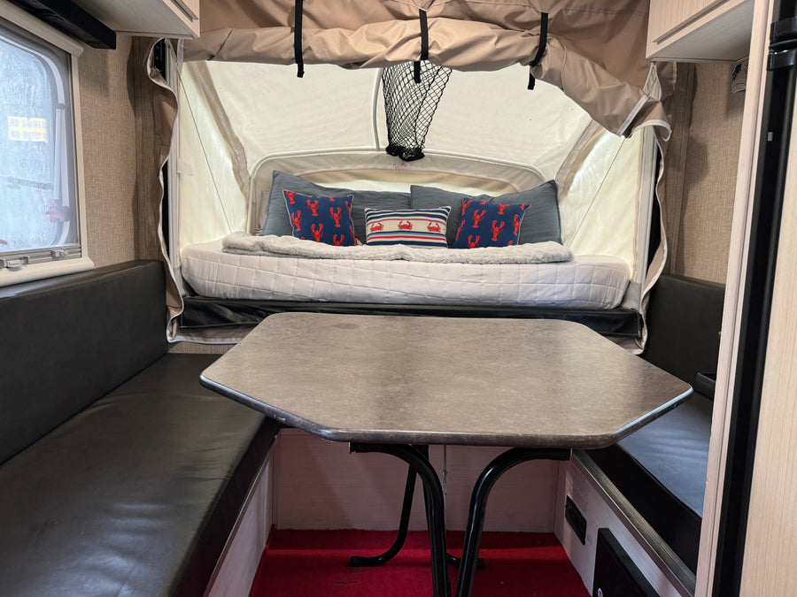 An RV rental's inviting interior: a central table, black booth seating, and a bed area with blue/red nautical pillows.