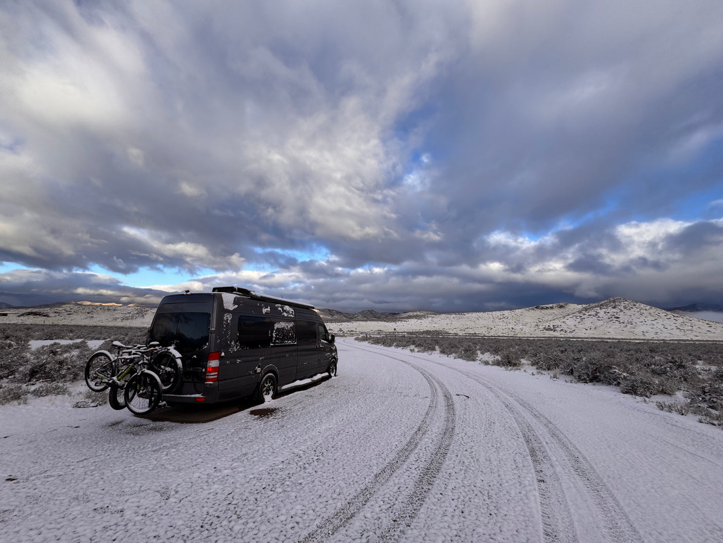 Dark gray RV rental with bikes on a snowy desert road. Dramatic winter clouds loom over snow-dusted hills.