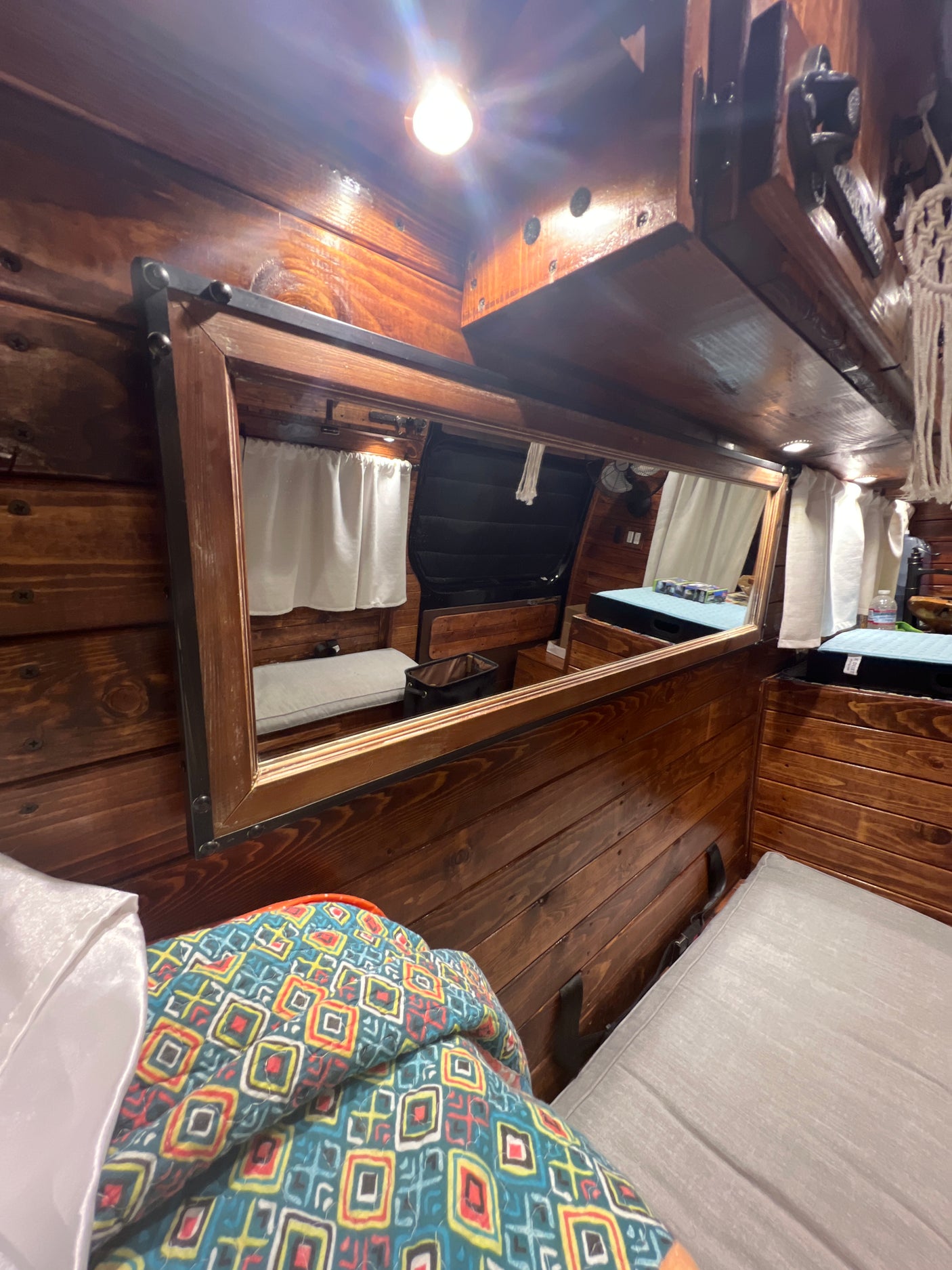 Inside an RV rental, a mirror reflects the cozy, wood-paneled interior featuring beds and a window with white curtains.