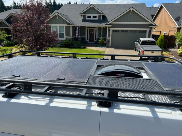 RV rental van's roof: black rack, solar panels, & MaxxAir fan. Ideal for mobile living, parked by a house.