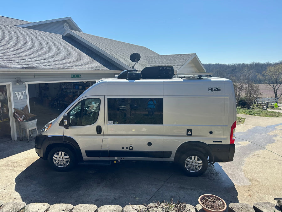 A silver RIZE camper van, perfect for an RV rental, is parked outside a home on a sunny day.